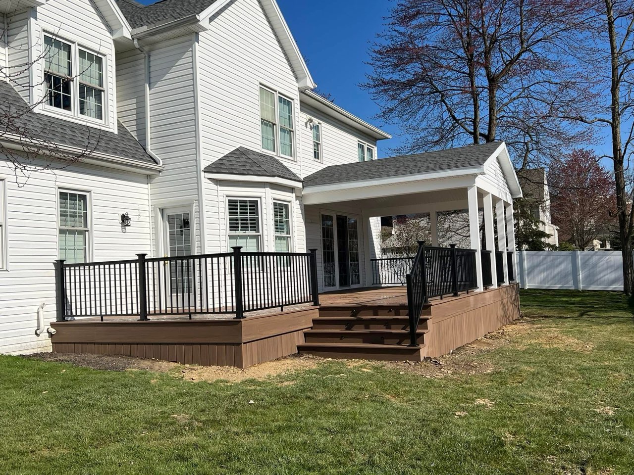 A white house boasts a large wooden deck crafted by expert deck builders, with sleek black railing and a covered section. The lush green grass surrounds the area, complemented by a nearby tree.