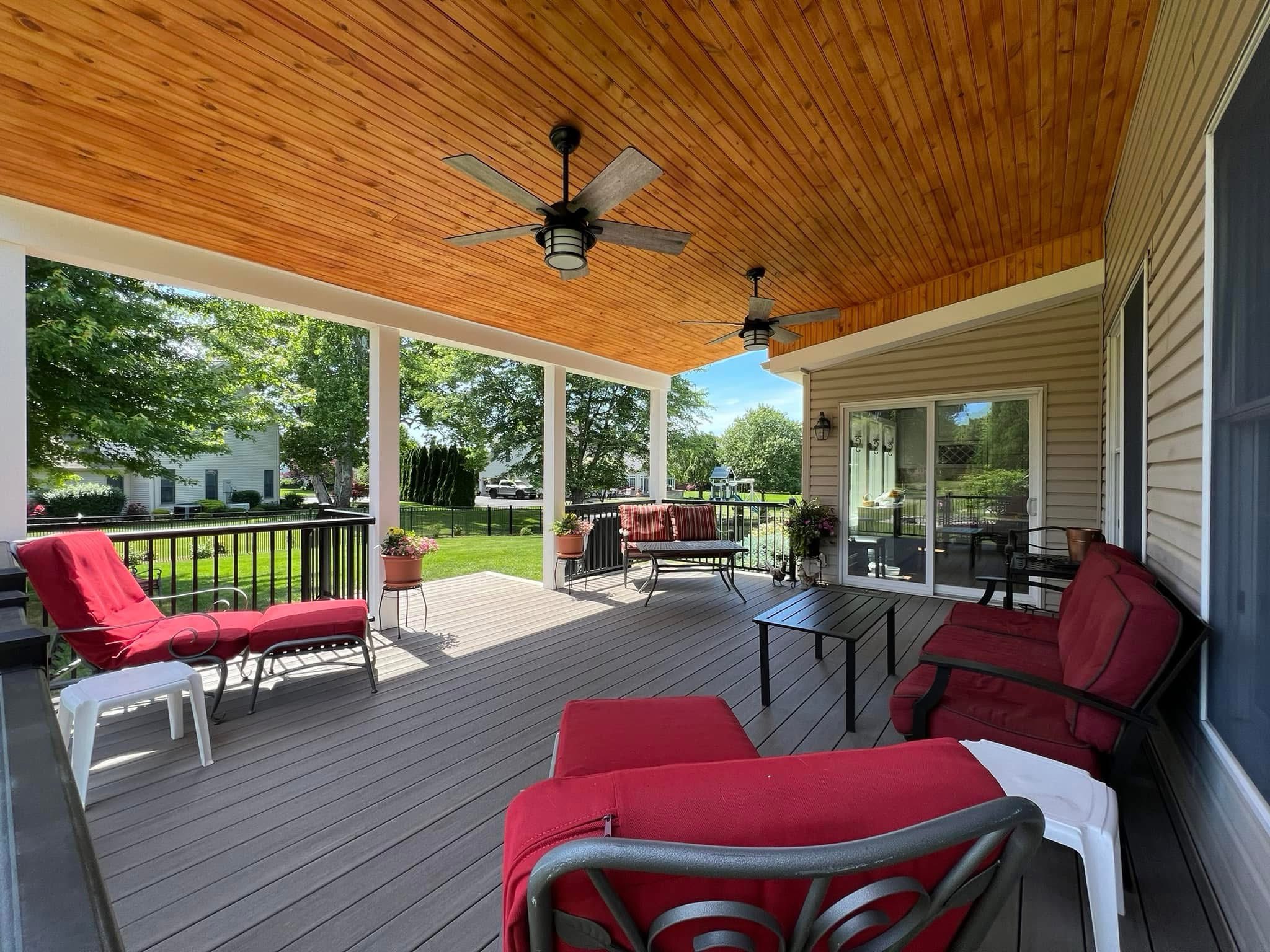 A covered porch with a wooden ceiling, ceiling fans, and red cushioned seating invites relaxation. Enjoy the table space while taking in views of greenery and houses, embodying the charm often found in well-designed covered decks.