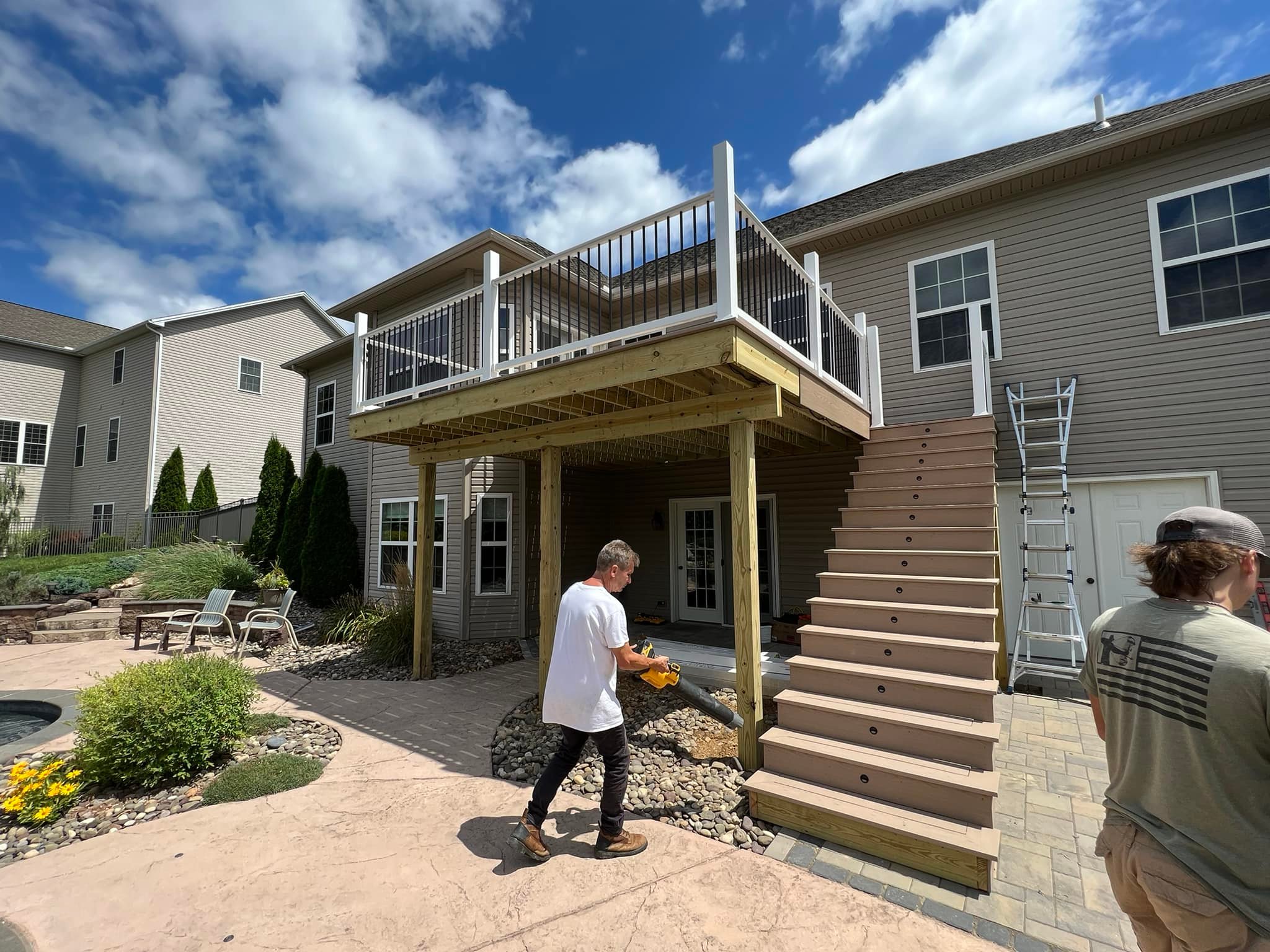 A person walks beneath a wooden deck attached to a house. Another person stands nearby. Outdoor furniture and a ladder are visible. The sky is partly cloudy.