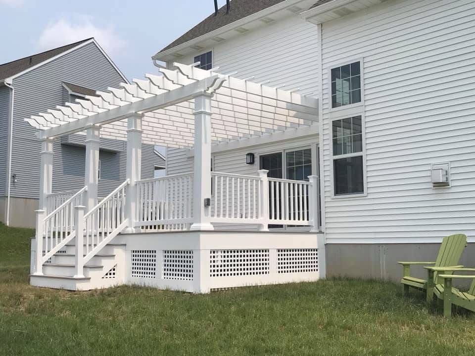 A white pergola, expertly crafted by skilled deck builders, is seamlessly attached to the house, with stairs descending gracefully to a grassy yard adorned with a charming green chair.