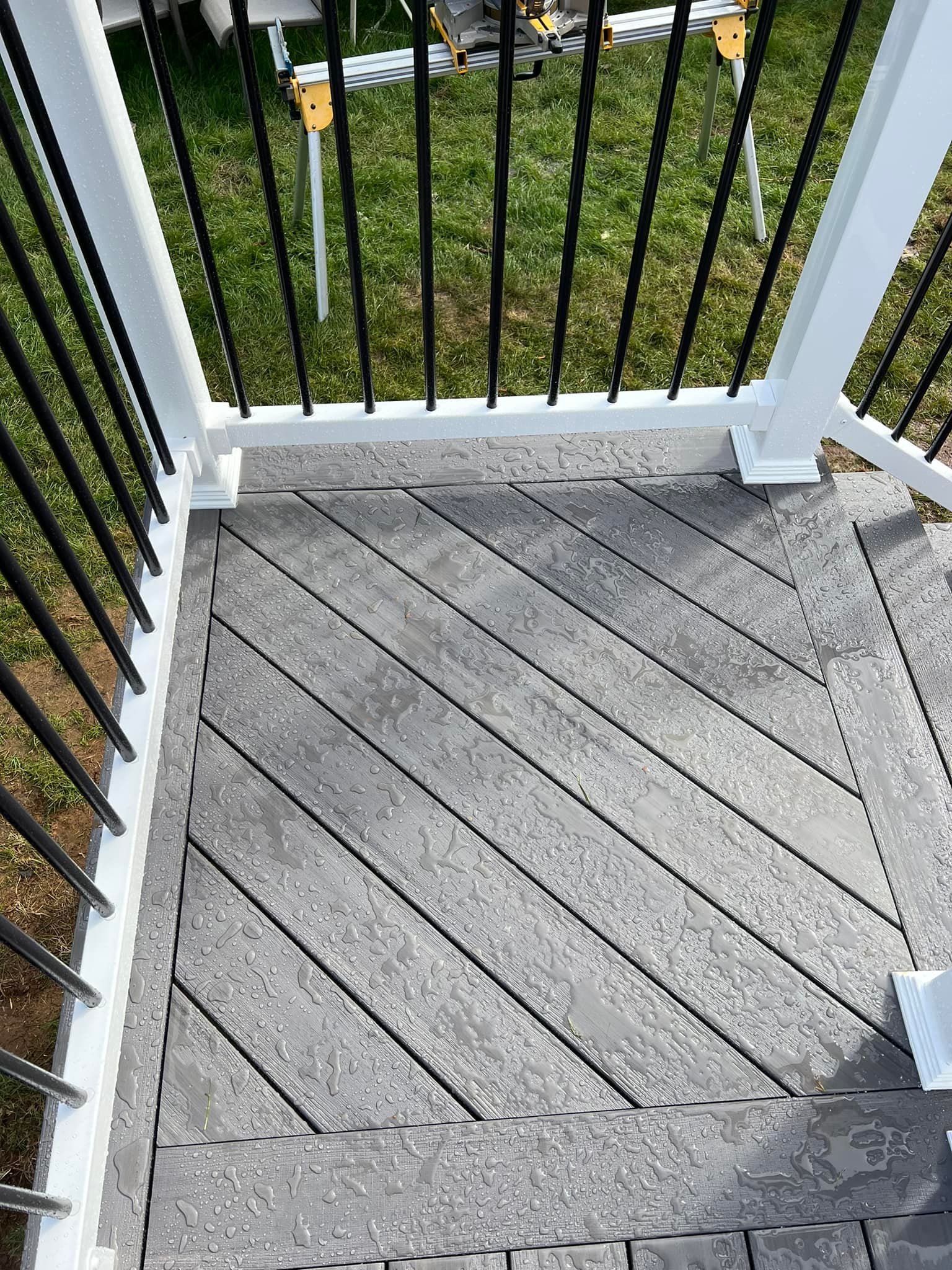 Wet gray deck flooring with raindrops, enclosed by black metal railings and white posts, adjacent to a grassy area with visible tools in the background.