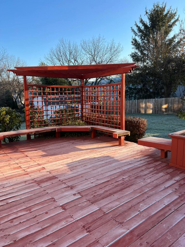 A wooden deck with integrated benches and a lattice pergola under a clear sky. The yard in the background includes trees and a wooden fence.