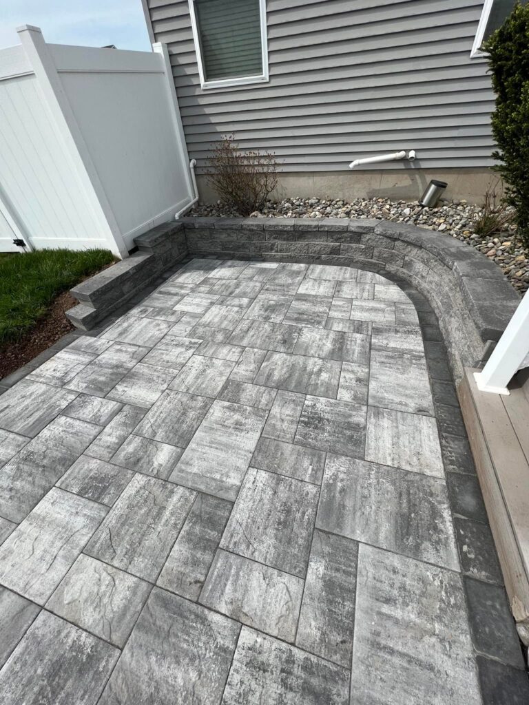 Gray stone patio with patterned tiles, surrounded by a white fence and a low stone wall, next to a house with gray siding.