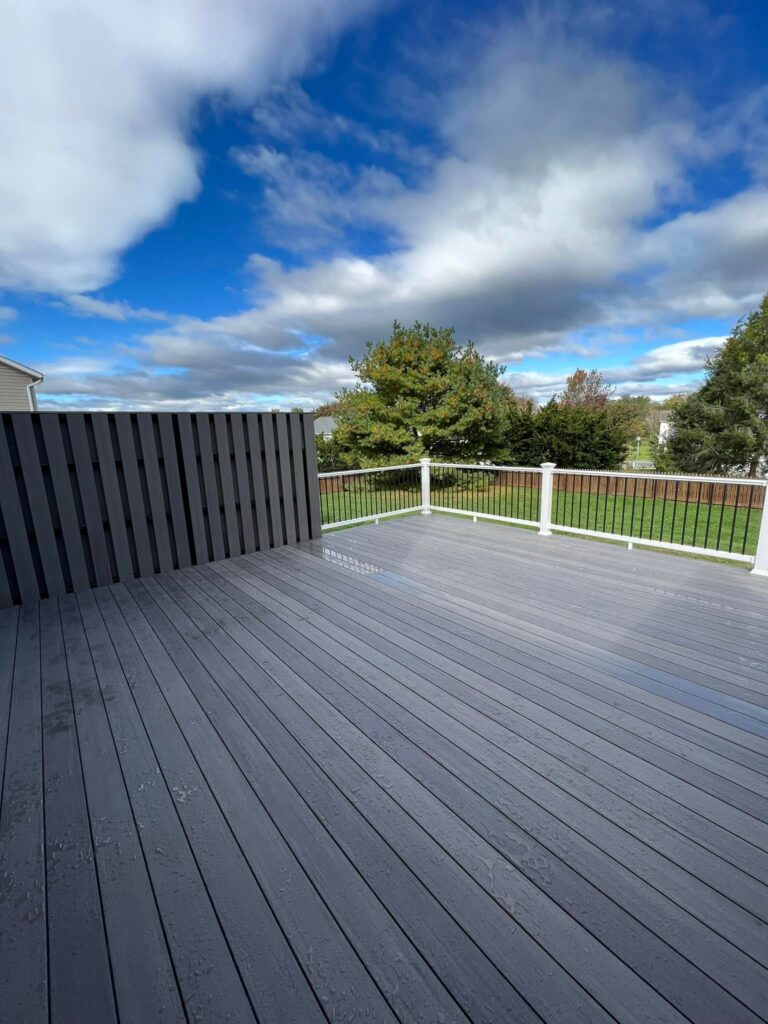 A gray wooden deck overlooking a grassy yard with trees under a partly cloudy sky.