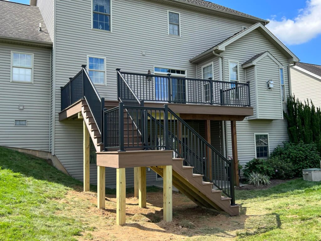 A two-story house with a newly built wooden deck and staircase, featuring black metal railings, in a backyard setting on a sunny day.