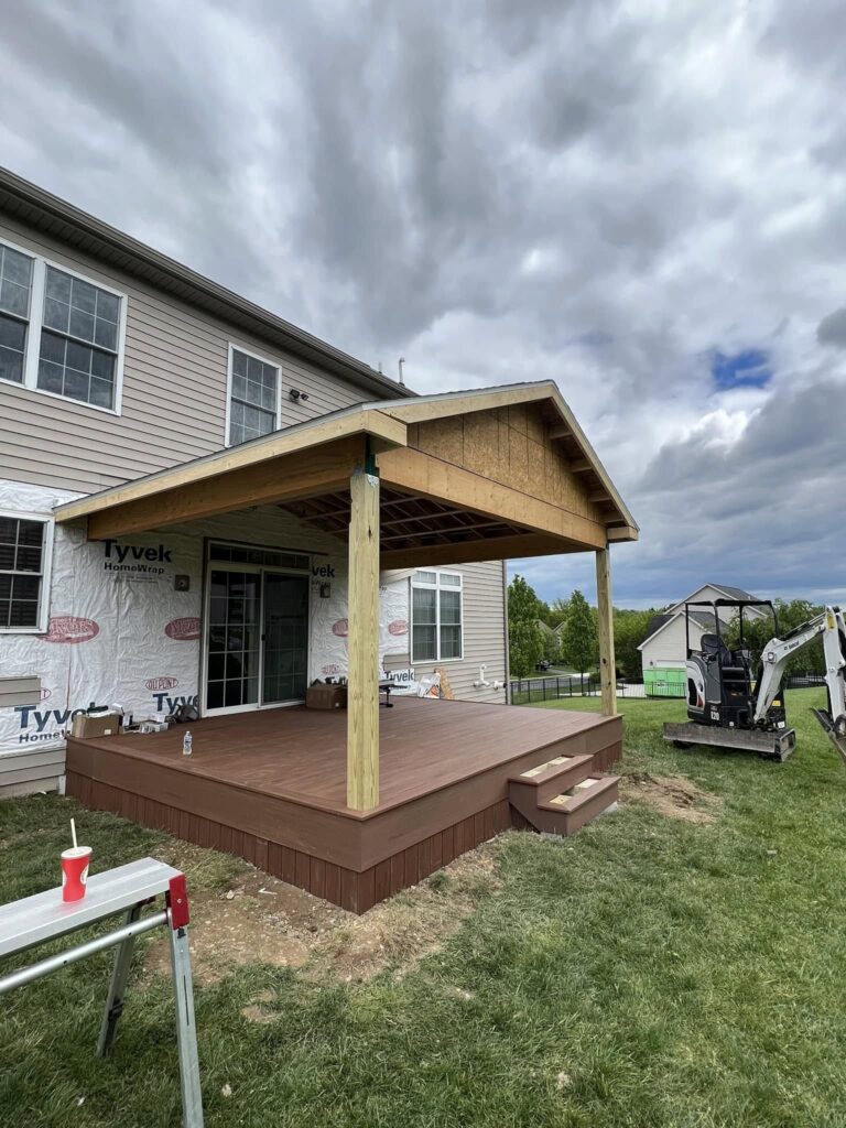 A partially constructed wooden deck with a roof is attached to the back of a house. Construction tools and equipment are visible in the surrounding yard.