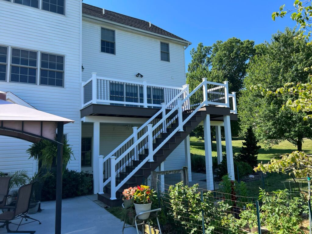 A white house with a raised wooden deck and stairs leading down to a patio. There are plants and trees in the yard.