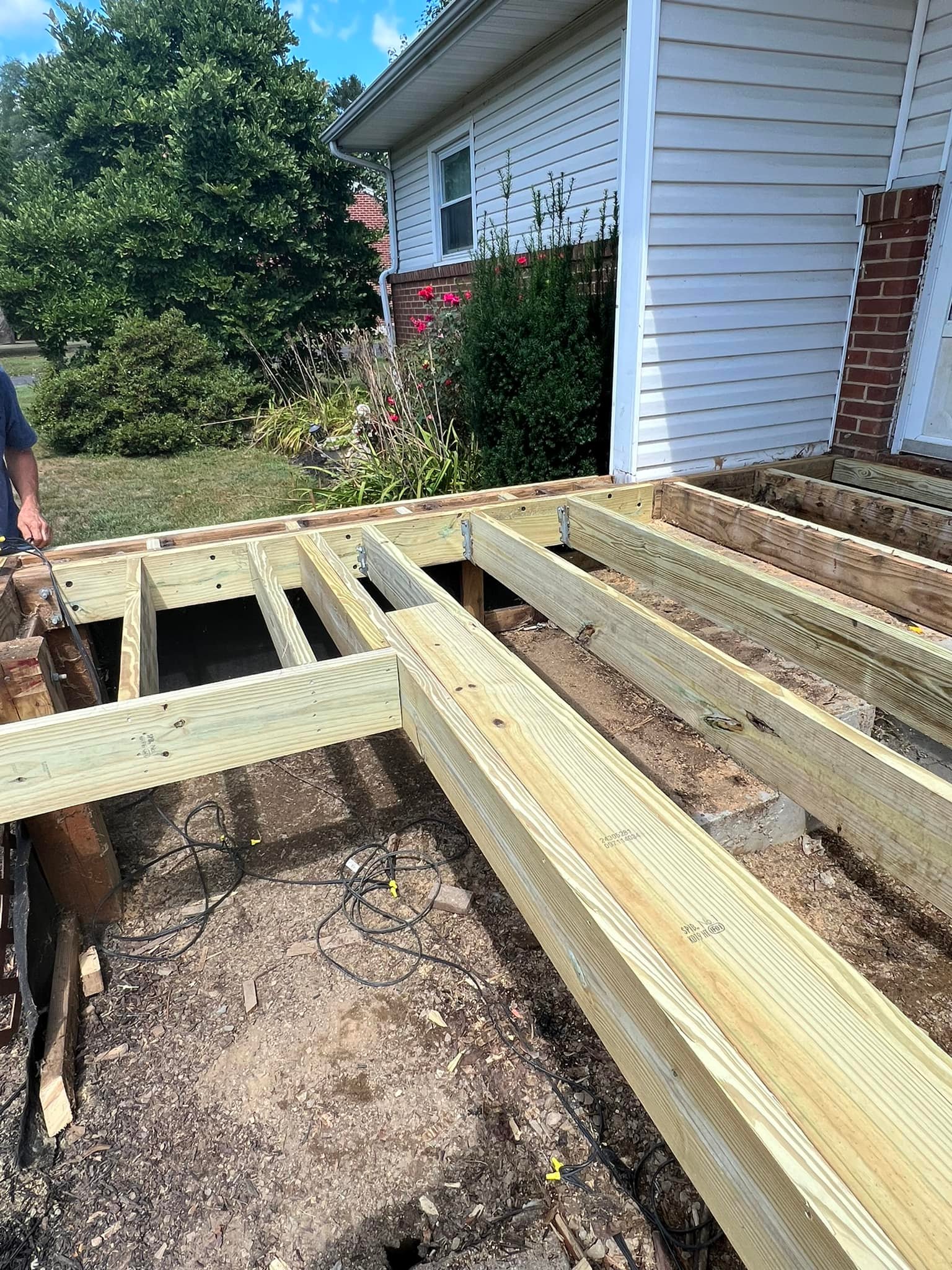 Partially constructed wooden deck attached to the side of a house, with visible beams and framework. Nearby are shrubs and a tree.