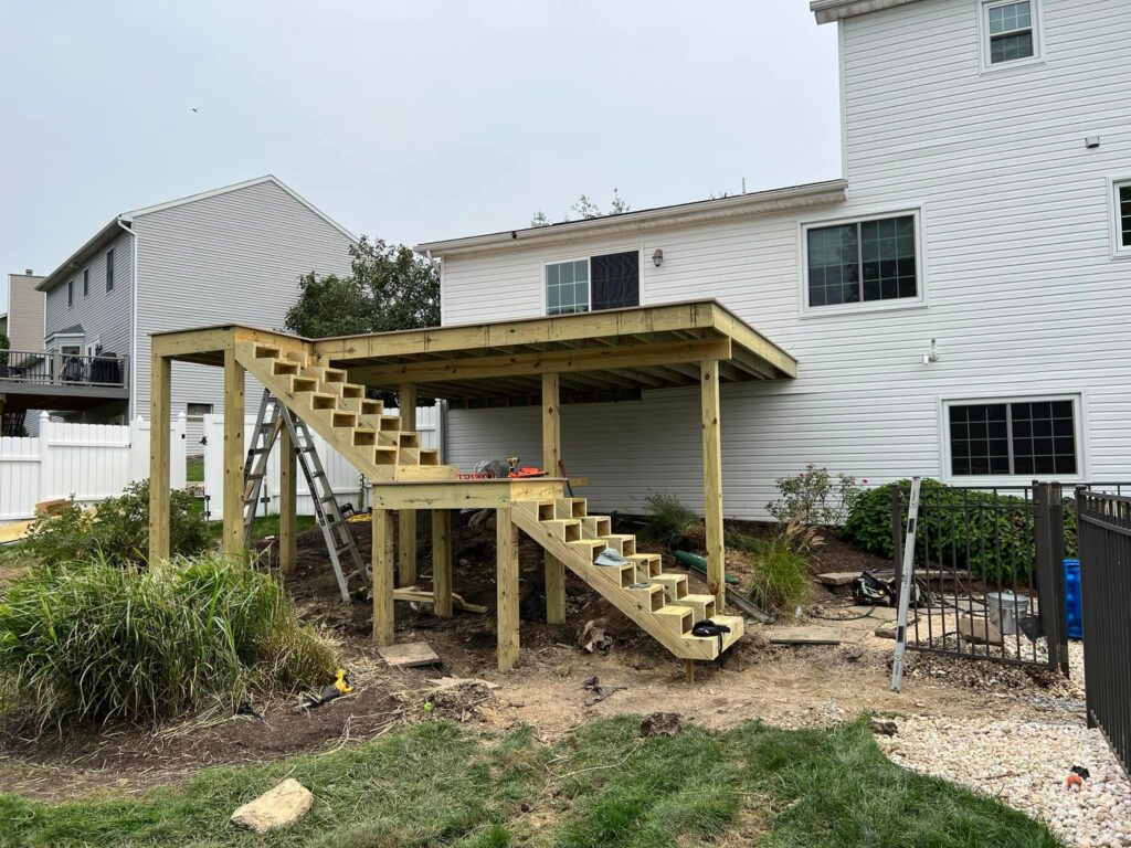 A partially constructed wooden deck with stairs is attached to a white house. Tools and materials are scattered around the area.